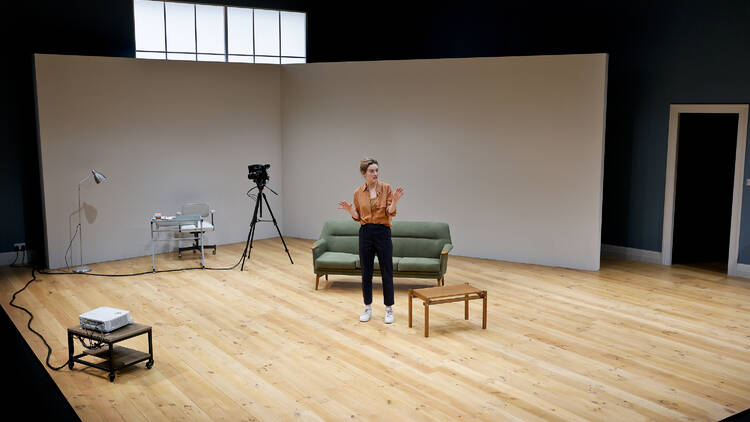 A woman stands on a stage with a couch, video camera and small coffee table.