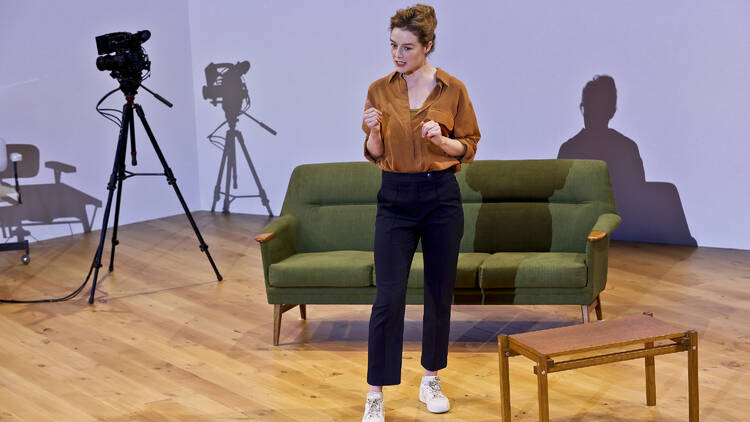 A woman stands on a stage in front of a green couch and next to a video camera.