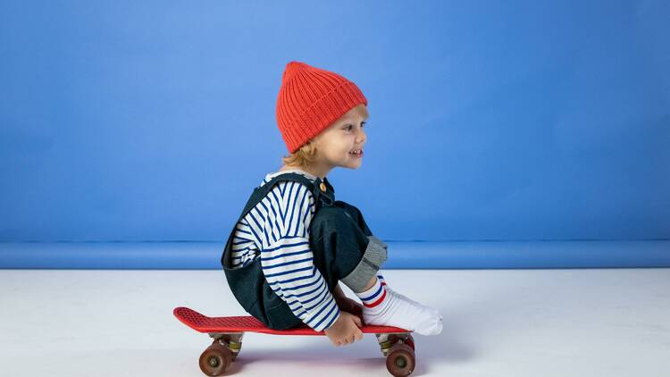 A young child wearing a red beanie and sitting on a skateboard.