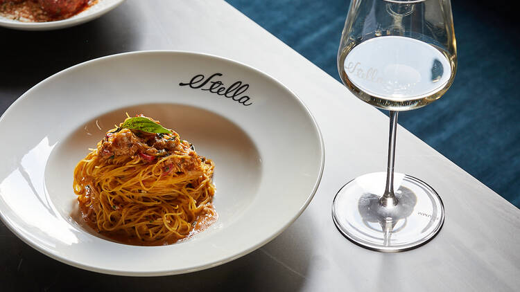 On a table there is a glass of white in next to a bowl of spaghetti and the bowl is branded with the word 'Stella'