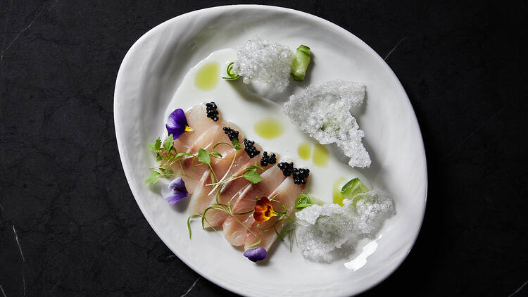 On a black table there is a white plate with kingfish slices, crispy crackers and garnishes