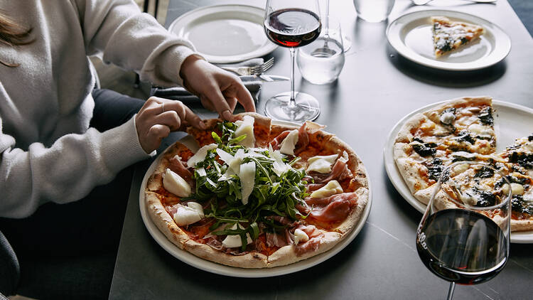 A person sitting at a table reaches for a slice of pizza which sits on the table next to glasses of red wine and another pizza