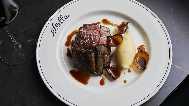 On a white plate with the words Stella printed on the plate there is sliced steak next to mashed potato