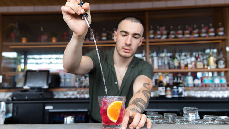 A bartender stands behind a bar and pours a spirit from above into a cocktail glass filled with pink liquid and an orange