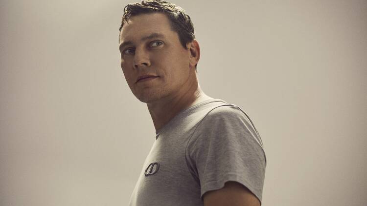 Tiësto in a grey t shirt looks to the left of the camera against a plain background.