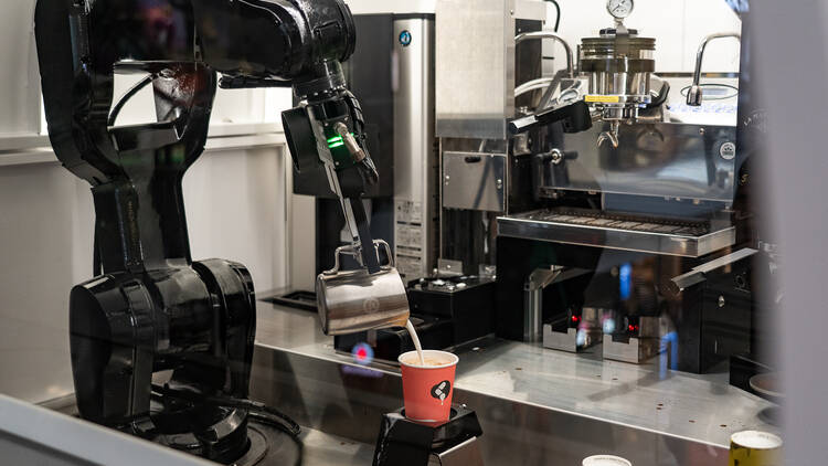 A robot pours milk from a silver jug into a take away coffee cup in front of a large coffee machine