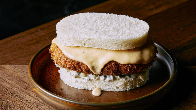 On a silver plate there is a sandwich made with white bread filled with crumbed fish topped with a mayonaisse sauce