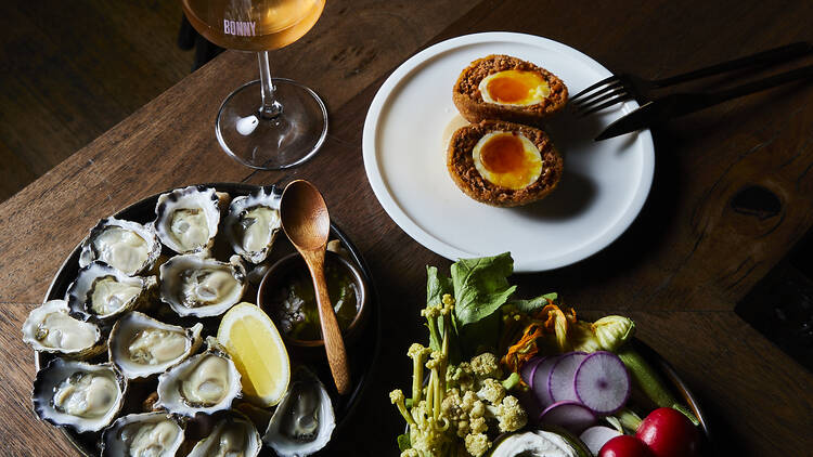 On a wooden table there is a glass of orange wine, a plate of oysters, a scotch egg and a plate of crudites