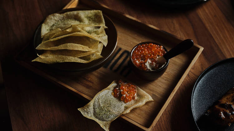 On a wooden board there is a bowl of crackers next to a small bowl of dip topped with roe being spooned onto a cracker