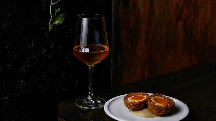 On a wooden table there is a glass of orange wine next to a plate with a scotch egg cut in hafl