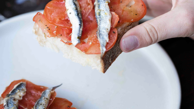 On A white plate there is a piece of bread topped with sliced topped and anchovy fillets, and someone picks up a half piece of the bread 