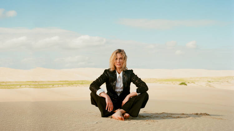 Delta Goodrem sits cross-legged on a sand dune while wearing a black suit and white shirt.