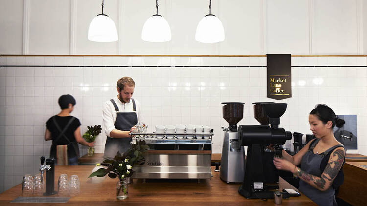 Inside Market Lane Coffee three barista work around several large coffee machines on a wooden bench above which there are low hung light fittings