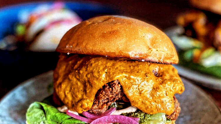 A burger sits on a blue plate which is filled with a chicken patty, lettuce, onions, and a butter chicken orange sauce