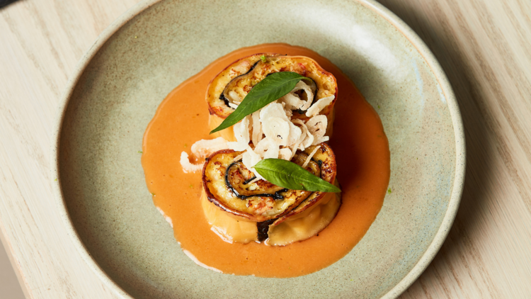 On a pastel blue green plate which is placed on a wooden table there are two large rolled pastas topped with a deep orange sauce