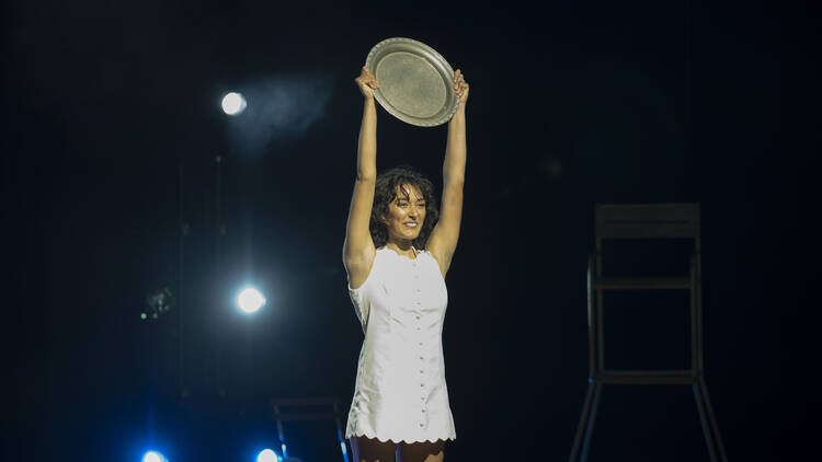 A woman in a white tennis dress holds a trophy up on stage.