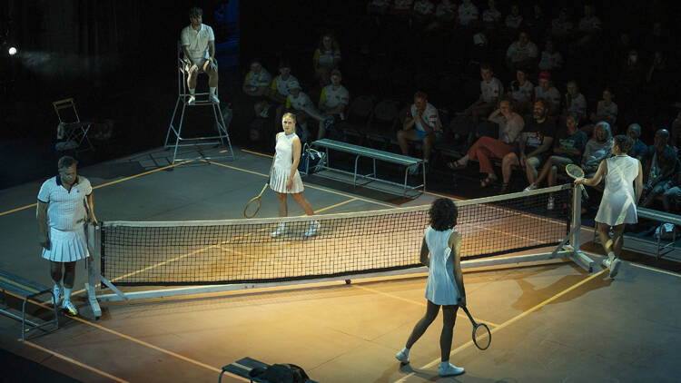 Four people in white tennis outfits stand on a mock tennis court on a stage.