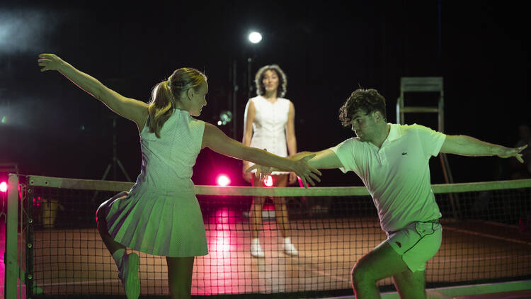 Three people in white tennis outfits pose on a mock tennis court on stage.