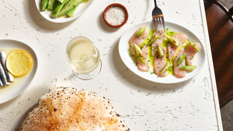 An image of a white table which has woodfired bread, a plate of cucumbers, sashimi and a glass of wine on top