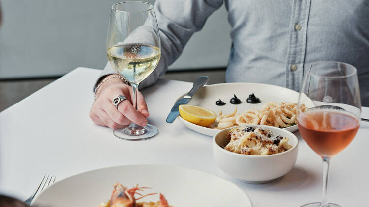 On a table there are several dishes of food including prawns and calamari, next to two glasses of wine, and you can see a person in a grey shirt holding onto one of the glasses of wine
