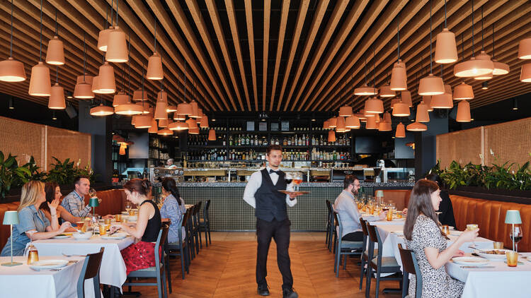 Inside the restaurant there are wooden floors, tables with white tableclothes, low hanging light fittings, and a waiter in a waistcoat walks through serving a cocktail