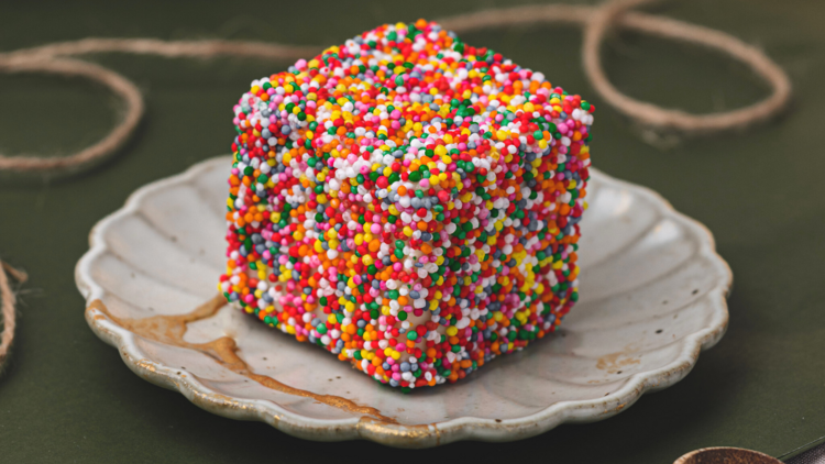 A square lamington sits on a porcelain plate which is covered in rainbow sprinkles