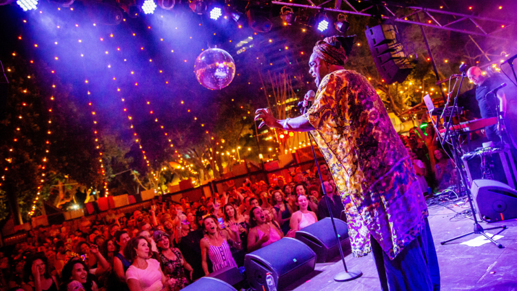 A performer in colourful attire stands on a glittery stage at night singing