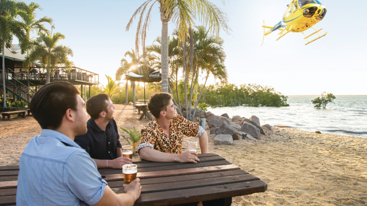 People sit at a table outside having sunset drinks looking at a helicopter overhead on Crab Claw Island in Darwin