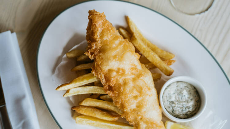 On a wooden table there is a plate of fried fish and chips with a lemon wedge and tartare sauce