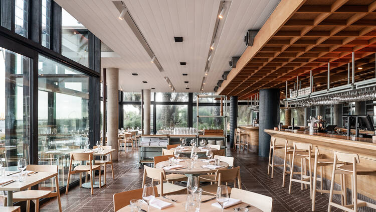 Inside the restaurant there are high ceilings and large waterfront windows, light wooden furniture and a long wooden bar