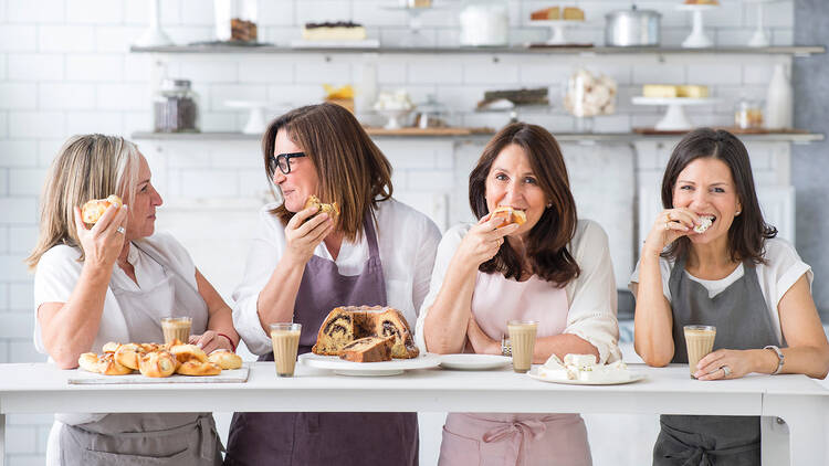 A group of four women stand at a bench eating cake and drinking coffee wearing aprons and smiling