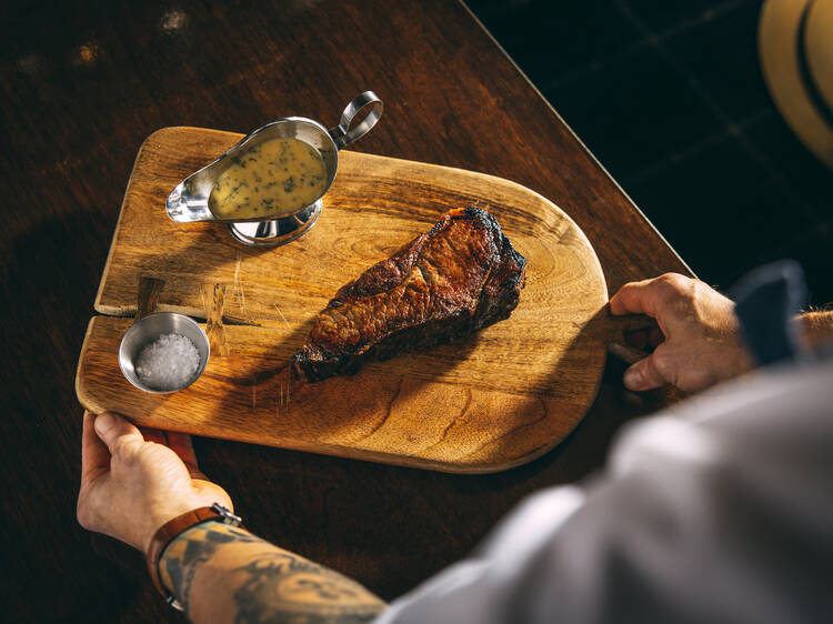 A pair of hands holding a wooden platter with a steak and sauce