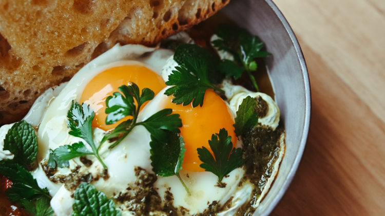 In a bowl there is two fried eggs with spices and herbs next to a crusty piece of bread