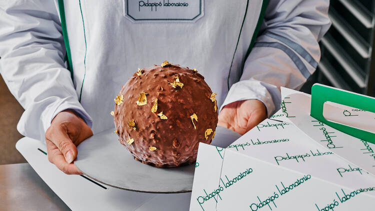 Someone in a Pidapipo apron holds a large cake that looks like a large Ferrero Rocher chocolate next to to a pidapipo box