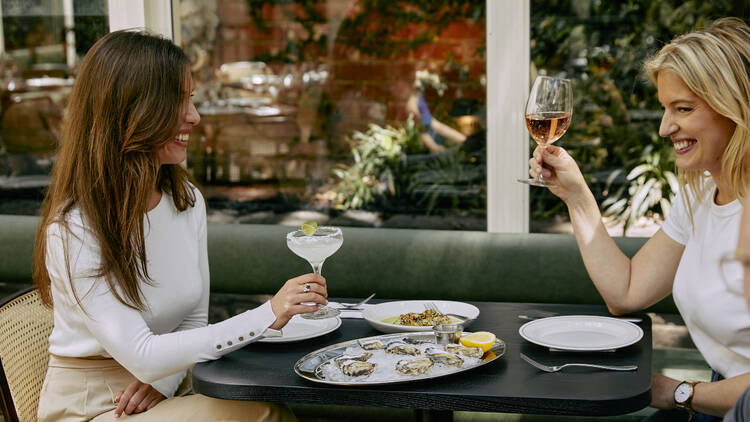 There is a small table next to a green couch with two women sitting at a table drinking cocktails with a plate of oysters