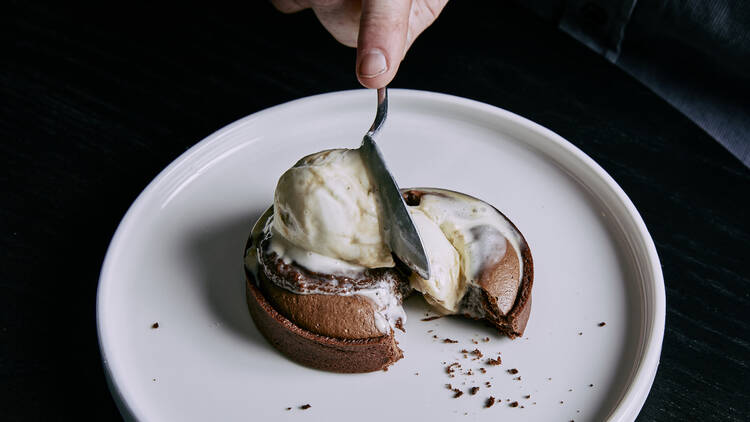 On a white plate there is chocolate fondant topped with ice cream with someone cutting into it