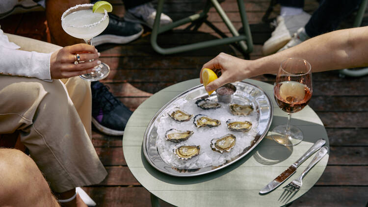 On a table outdoors there is a large tray of oysters on ice next to a glass of rose and someone squeezes a lemon onto an oyster