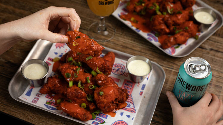 There is a silver tray piled high with chicken wings next to small bowls of dipping sauce with someone holding a chicken wing and a beer can in the other hand