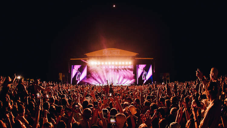 An outdoor music festival at night, with a lit-up stage and crowds of people.