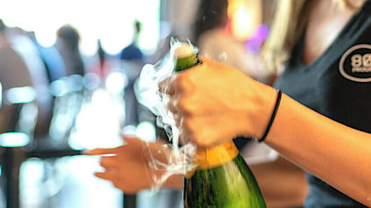 A person pops a bottle of champagne at 80 Proof
