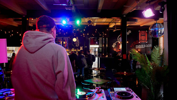 There is a DJ standing behind DJ decks in a dark room with colourful lights in front of a dance floor