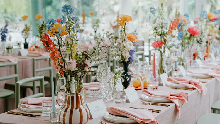 There are long tables with pink tableclothes topped with many different vases of colourful flowers