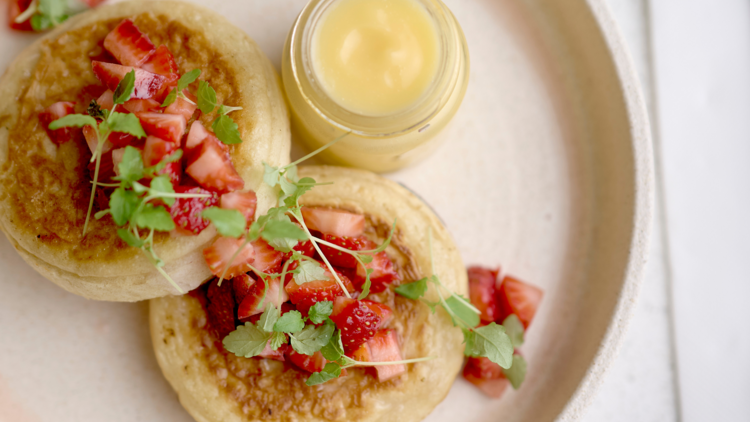 There is a white plate with two crumpets topped with strawberries and a small jar of lemon curd