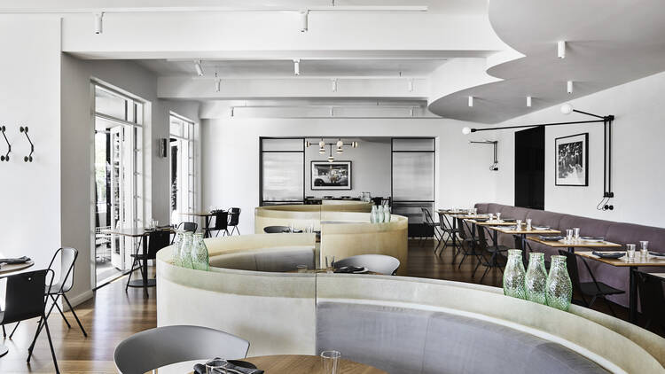 Inside the Prince Dining Room there is a curved structure around the banquette couches, white walls and ceilings and wooden tables