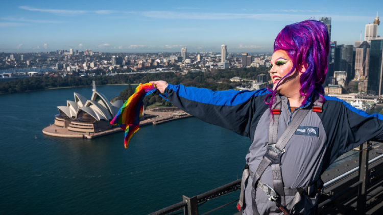A drag queen holding a rainbow flag stands on top of the Sydney Harbour Bridge