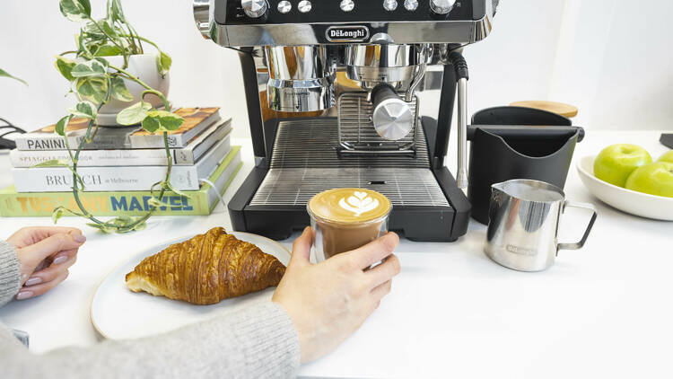 An espresso machine, cup of coffee and a croissant on a table.