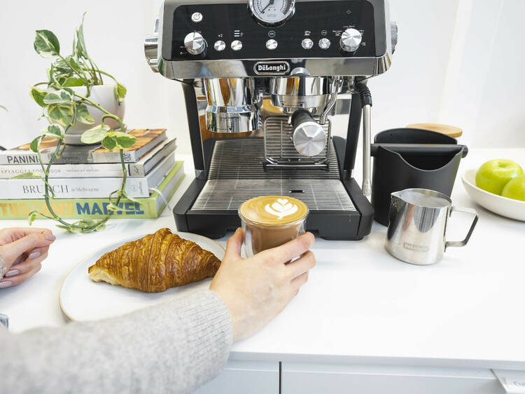 An espresso machine, cup of coffee and a croissant on a table.