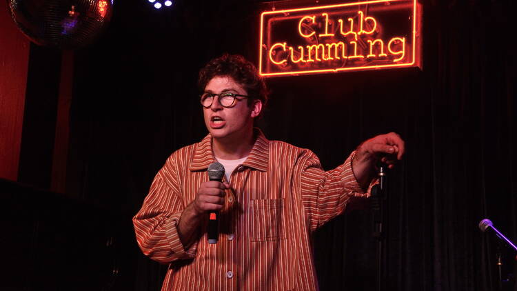 A performer holds a mic in front of a neon sign reading Club Cumming.