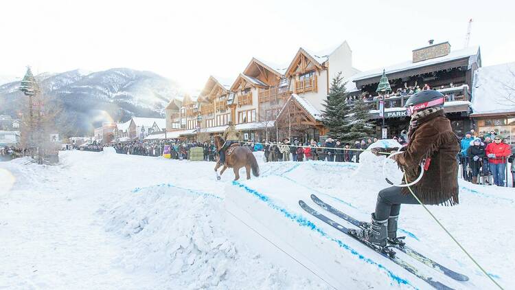 A horse pulls a skier up a snow ramp as gathered spectators enjoy a wintry day