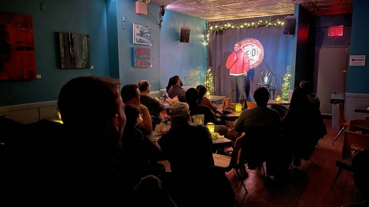 A performer in a red hoodie stands on stage at QED Astoria. 